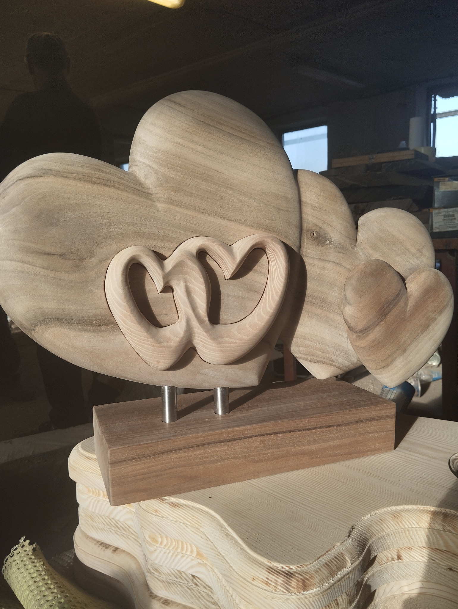 Dekoracyjna rzeźba z drewna przedstawiająca trzy serca na metalowych nóżkach, umieszczona na drewnianej podstawie; w tle warsztat stolarski.