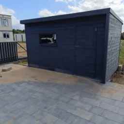 Ciemnoszara, drewniana konstrukcja z oknem i drzwiami, prawdopodobnie domek narzędziowy, ustawiona na szarej kostce brukowej, z widocznym fragmentem ogrodzenia i budynkiem w tle.