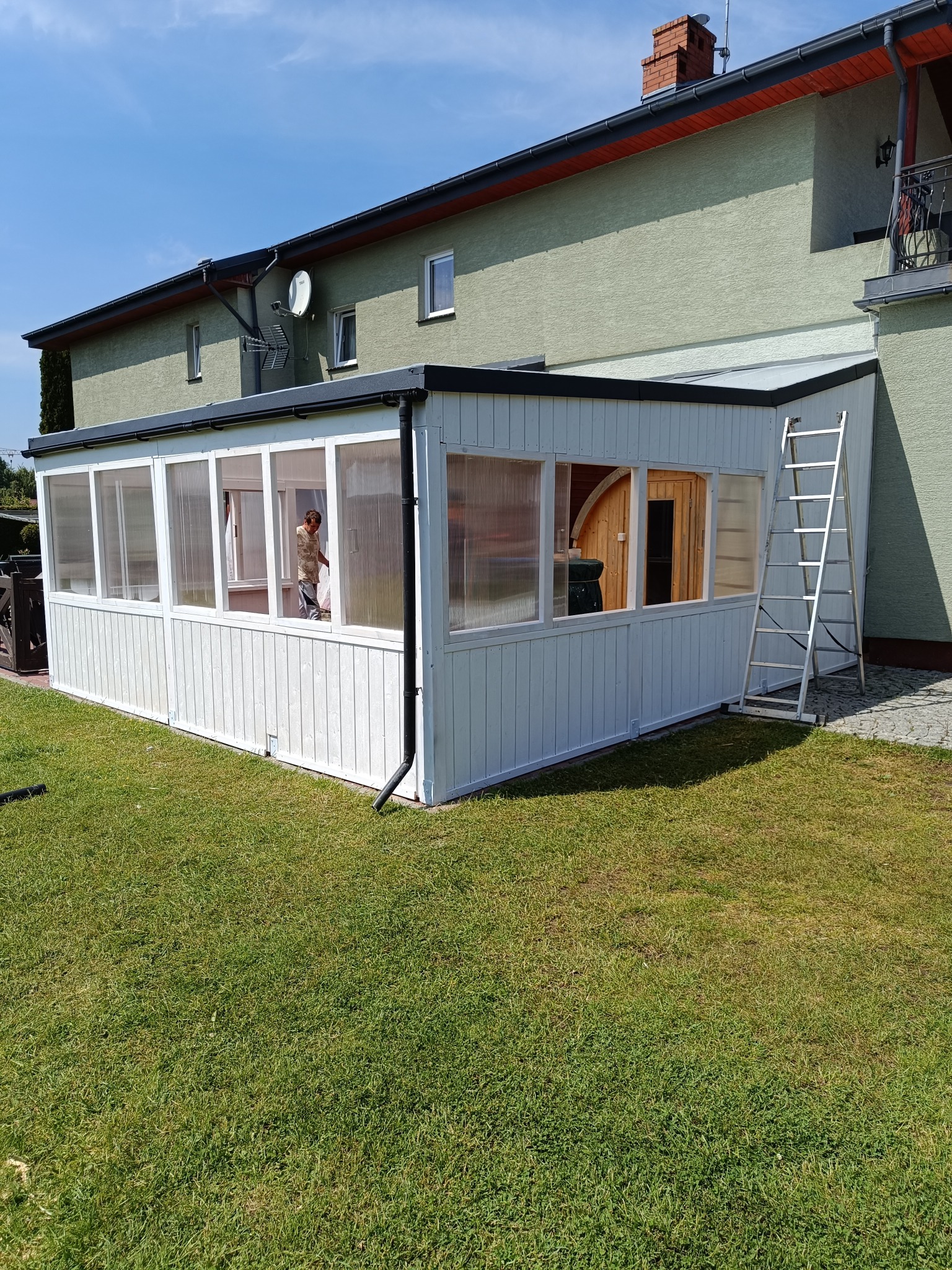 Biała konstrukcja z przeszkleniami, prawdopodobnie weranda lub oranżeria, przylegająca do domu z zieloną elewacją. Wewnątrz widoczna osoba oraz fragment sauny. Do ściany oparta aluminiowa drabina.