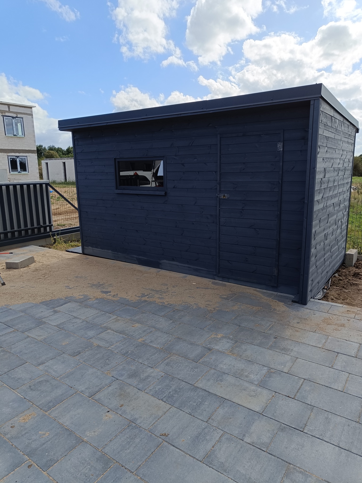 Ciemnoszara, drewniana konstrukcja z oknem i drzwiami, prawdopodobnie domek narzędziowy, ustawiona na szarej kostce brukowej, z widocznym fragmentem ogrodzenia i budynkiem w tle.