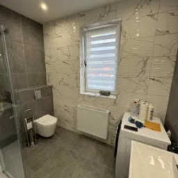 Nowoczesna łazienka z szarymi i białymi płytkami imitującymi marmur, kabiną prysznicową i oknem z żaluzjami. Umywalka częściowo widoczna na pierwszym planie.