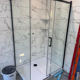 Andrzej Dobrovolskyi - Kabina prysznicowa z czarnymi profilami i półkami w wykończonej łazience z marmurowymi płytkami. Widoczne narzędzia i akcesoria montażowe.