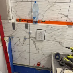 Montaż płytek imitujących marmur w łazience, widoczne narzędzia glazurnicze, poziomica i butelka wody. Prace wykończeniowe w trakcie realizacji.