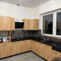 Andrzej Dobrovolskyi - Nowoczesna kuchnia z drewnianymi szafkami i czarnym blatem, okapem oraz płytą gazową. Widoczne akcesoria montażowe na półce. Szare płytki na ścianie i podłodze.