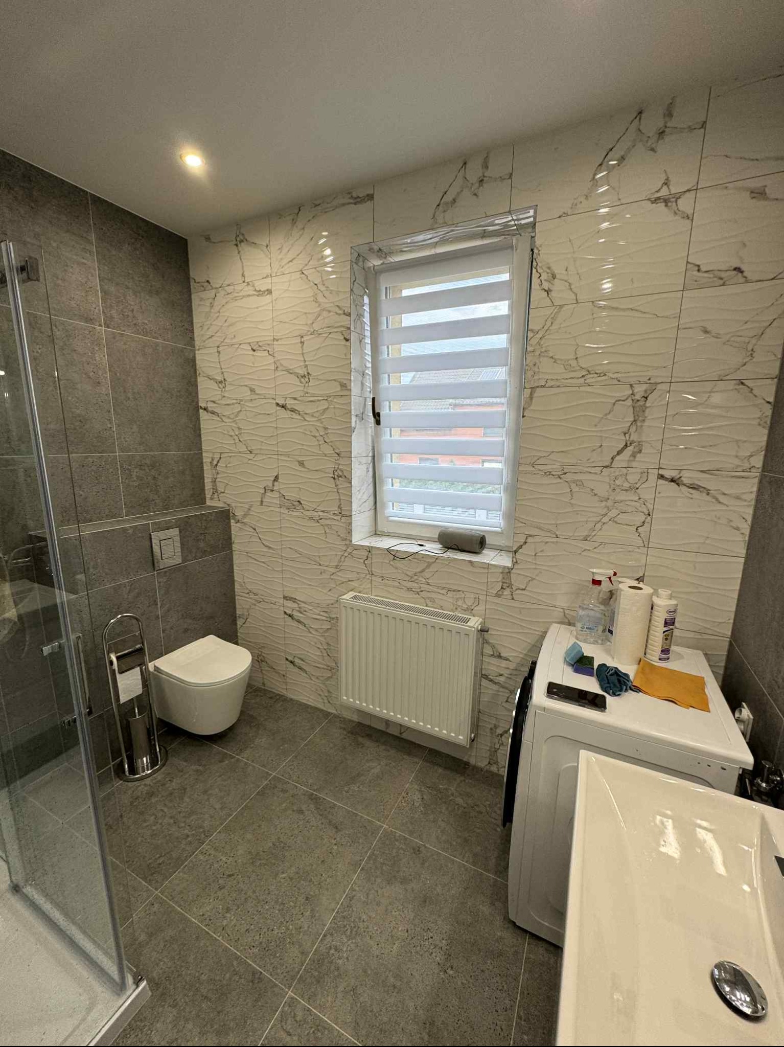 Nowoczesna łazienka z szarymi i białymi płytkami imitującymi marmur, kabiną prysznicową i oknem z żaluzjami. Umywalka częściowo widoczna na pierwszym planie.