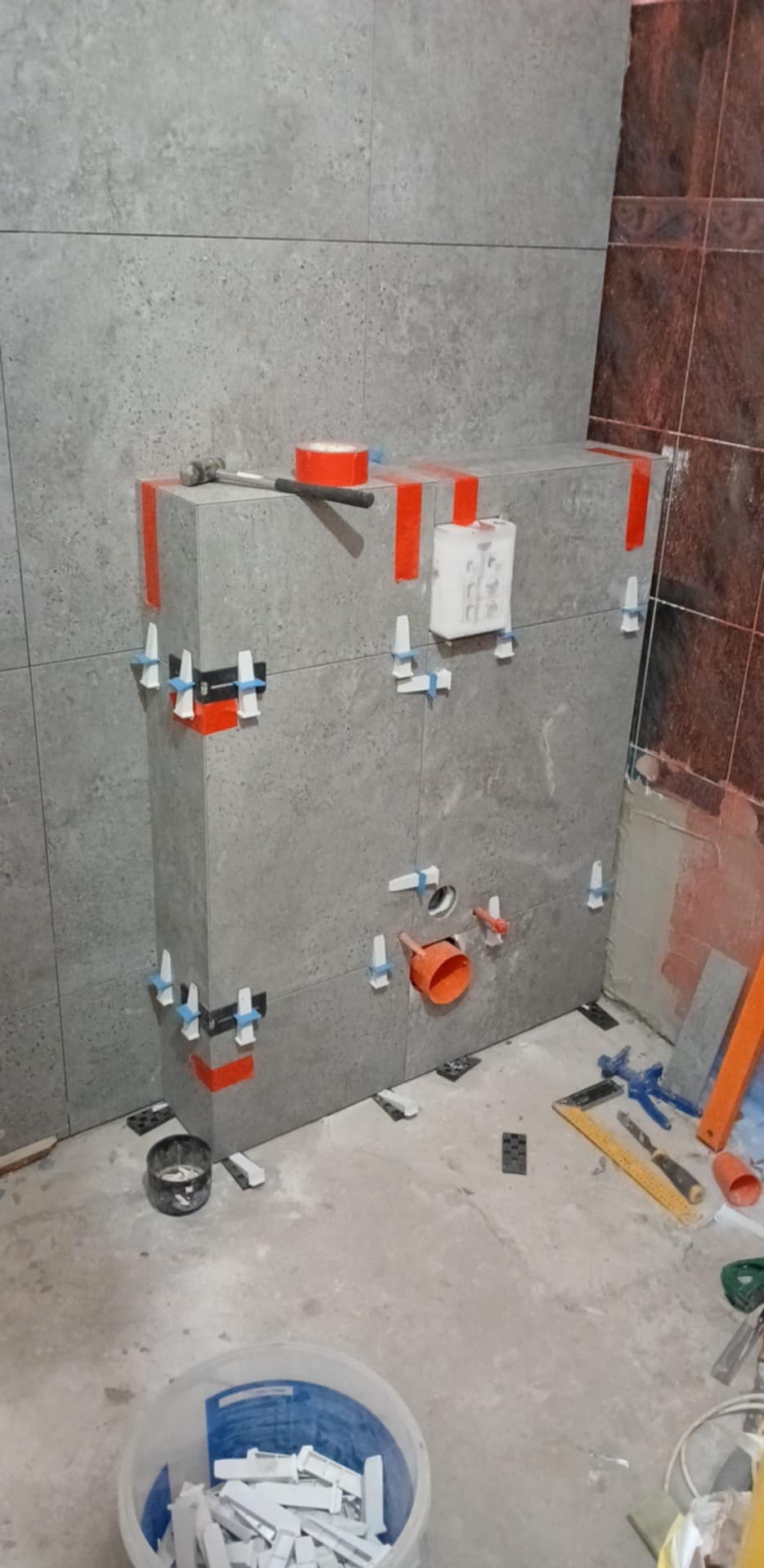 Montaż szarych płytek w łazience, widoczne narzędzia glazurnicze i poziomica. Częściowo wykończona ściana z instalacją. Na podłodze wiadro z krzyżykami dystansowymi.