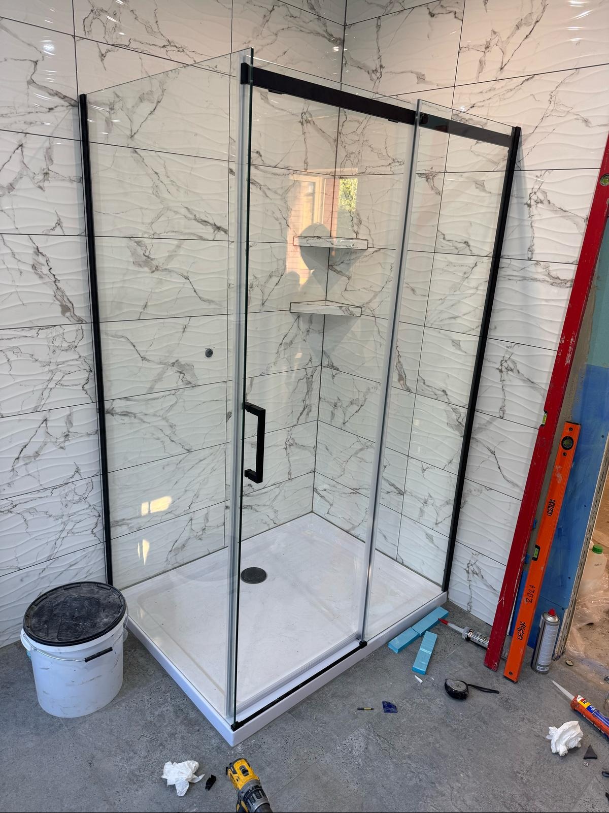 Kabina prysznicowa z czarnymi profilami i marmurowymi płytkami w łazience. Widoczne narzędzia i poziomica, co sugeruje etap wykończenia.