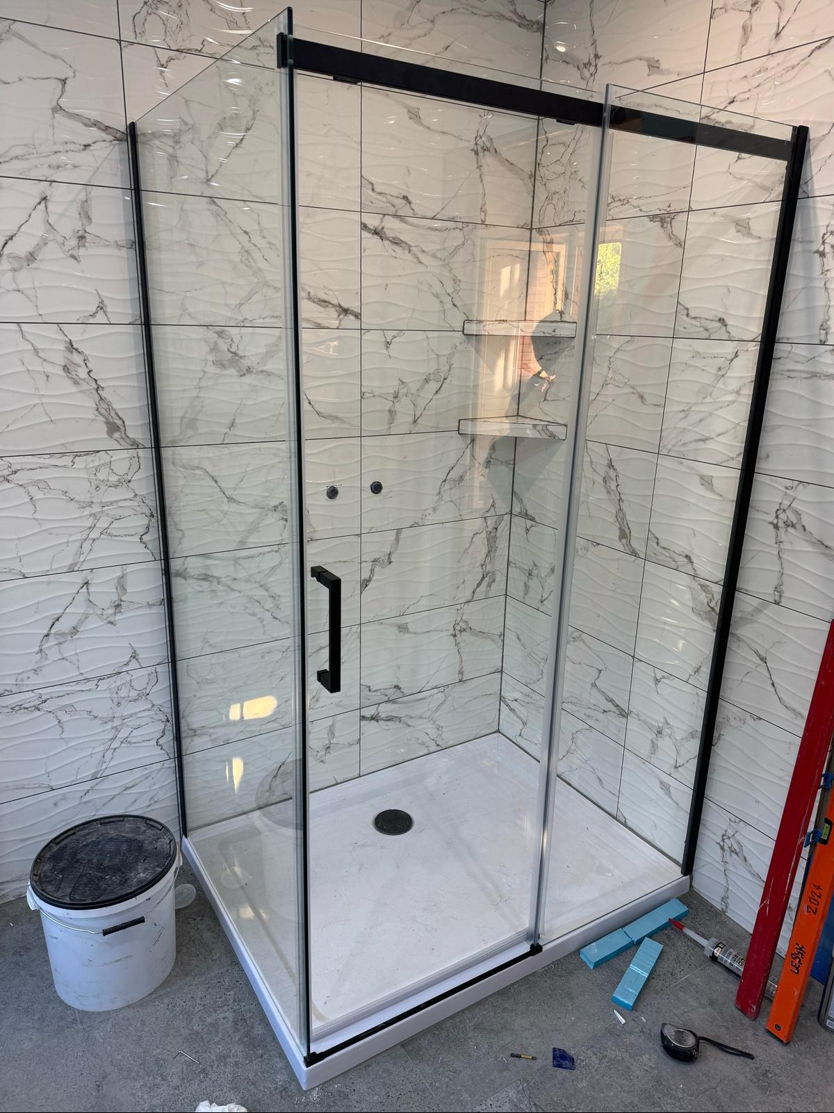 Nowoczesna kabina prysznicowa z czarnymi profilami i marmurowymi płytkami na ścianach. Widoczne narzędzia i wiadro, sugerujące świeży montaż.