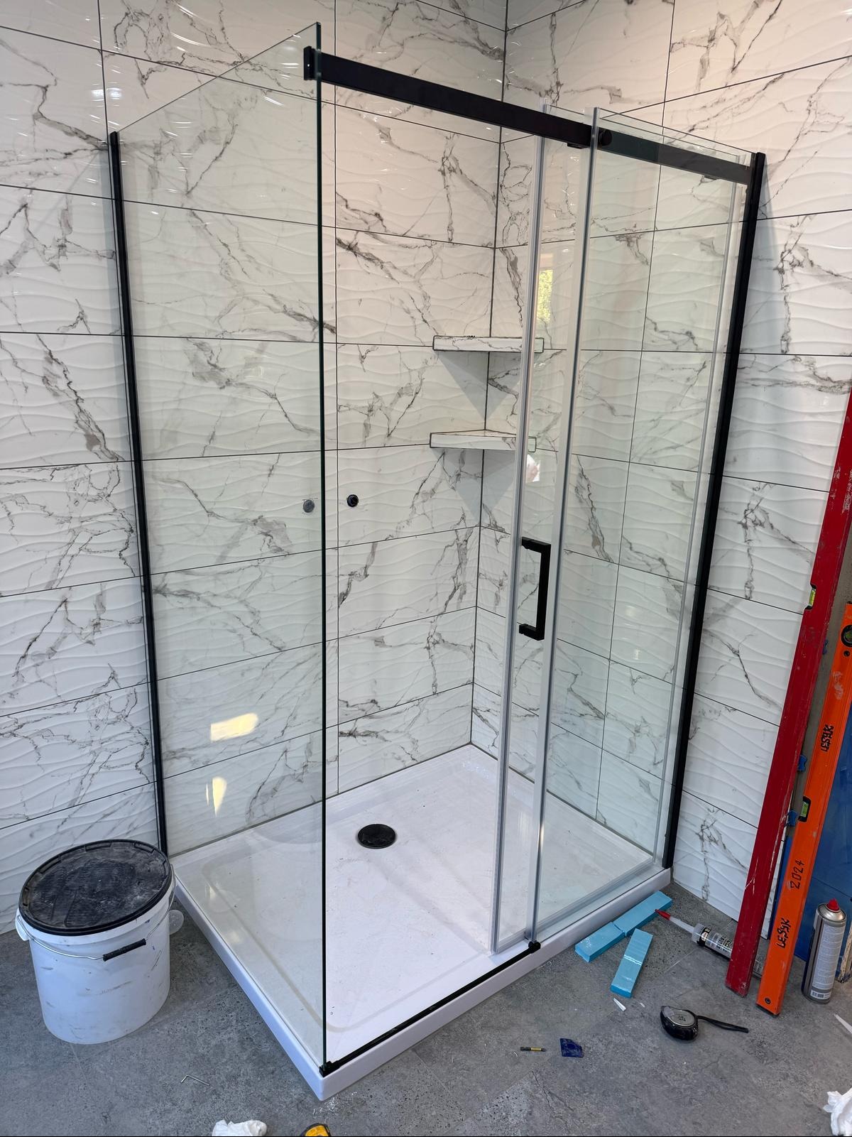 Kabina prysznicowa z czarnymi profilami i półkami w wykończonej łazience z marmurowymi płytkami. Widoczne narzędzia i akcesoria montażowe.
