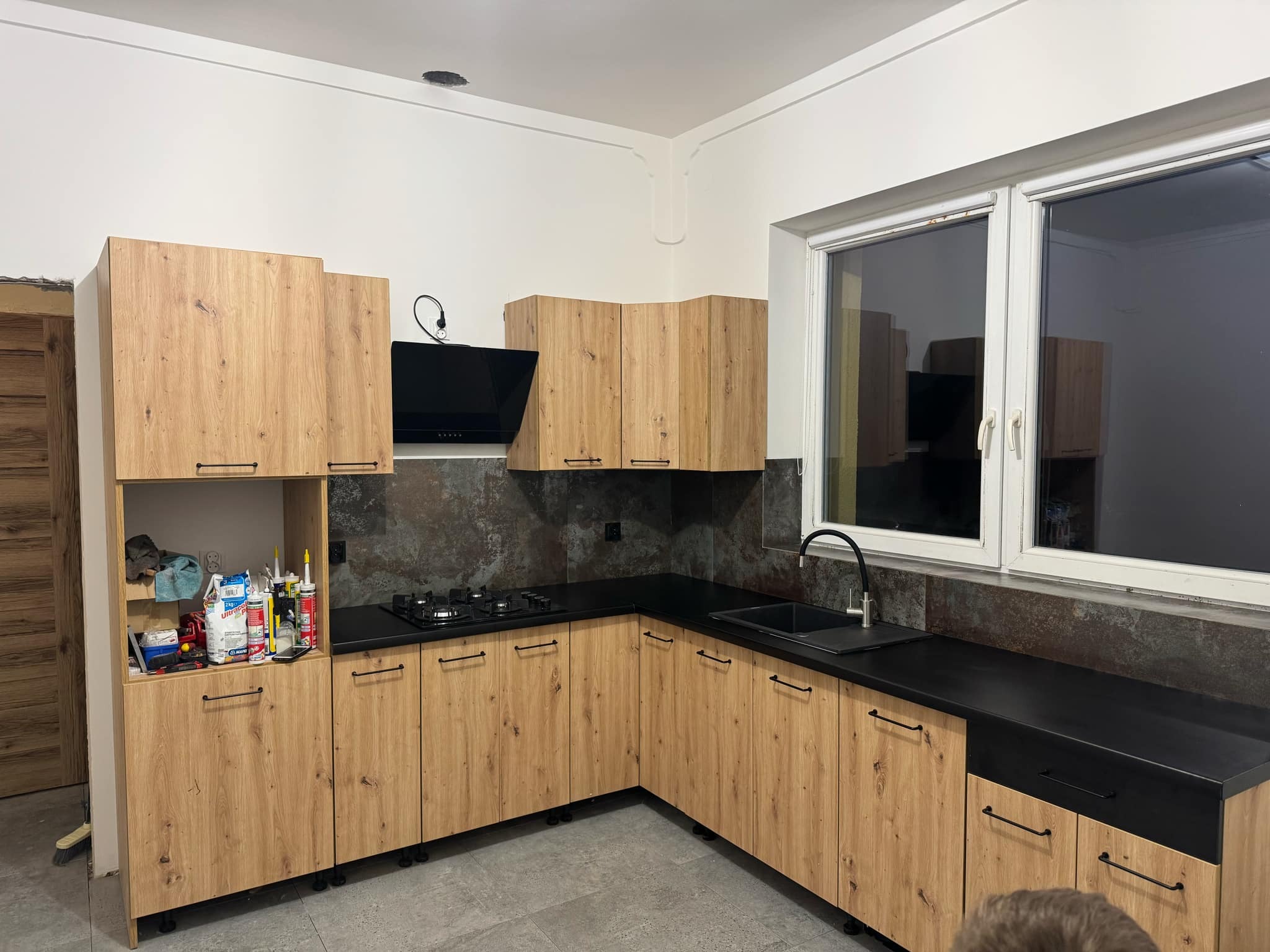 Nowoczesna kuchnia z drewnianymi szafkami i czarnym blatem, okapem oraz płytą gazową. Widoczne akcesoria montażowe na półce. Szare płytki na ścianie i podłodze.