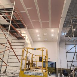 Konrad Rojek - Montaż płyt na suficie w dużym pomieszczeniu, widoczne rusztowania i podnośnik, szpachlowane łączenia płyt, w tle oświetlenie sufitowe