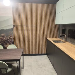 Konrad Rojek - Nowoczesna kuchnia z drewnianą ścianą z lameli, blatem w kolorze jasnego drewna i minimalistycznymi, jasnymi szafkami, uzupełniona o stół z ciemnego drewna i zielone krzesła.