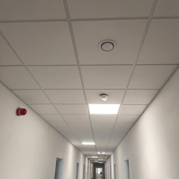 Konrad Rojek - Długi korytarz biurowy w trakcie wykańczania, widoczne białe ściany, szara podłoga, sufit kasetonowy z oświetleniem i czujnikiem dymu, czerwony alarm pożarowy na ścianie.