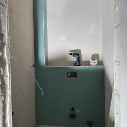 Konrad Rojek - Wnętrze łazienki w trakcie zabudowy: zielone płyty na stelażu, widoczne otwory na instalacje, wkrętarka akumulatorowa Makita i opakowanie wkrętów na półce, surowe ściany w tle.