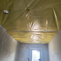 Konrad Rojek - Pokój w trakcie prac wykończeniowych: surowy tynk na ścianach, zabezpieczony folią sufit z wystającymi przewodami, drabina oparta o ścianę pod oknem.