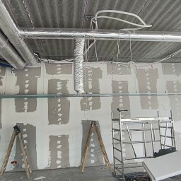 Konrad Rojek - Wnętrze w trakcie prac wykończeniowych: ściany z płyt karton-gips, częściowo zaszpachlowane, widoczne rury wentylacyjne pod sufitem, drabiny i rusztowanie.