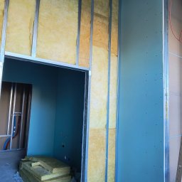 Konrad Rojek - Wnętrze w trakcie prac wykończeniowych: metalowy stelaż, wełna mineralna w ścianie, częściowo zamontowana płyta na ścianie, widoczne przewody instalacji.