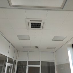 Konrad Rojek - Kwadratowy panel sufitowy z kratką wentylacyjną w biurze, widok z dołu, białe ściany i okna z czarnymi ramami.