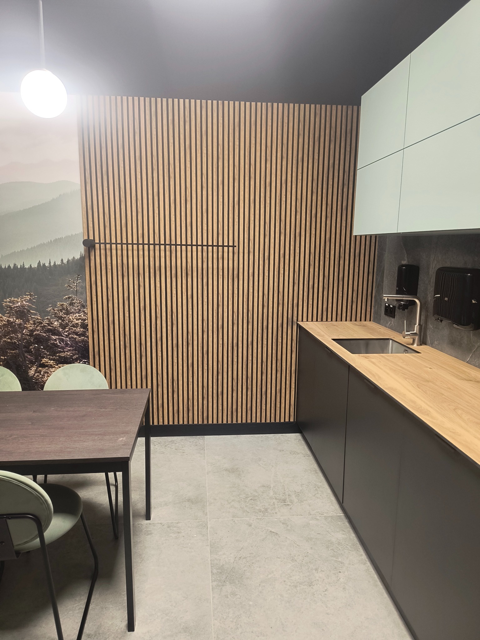 Nowoczesna kuchnia z drewnianą ścianą z lameli, blatem w kolorze jasnego drewna i minimalistycznymi, jasnymi szafkami, uzupełniona o stół z ciemnego drewna i zielone krzesła.