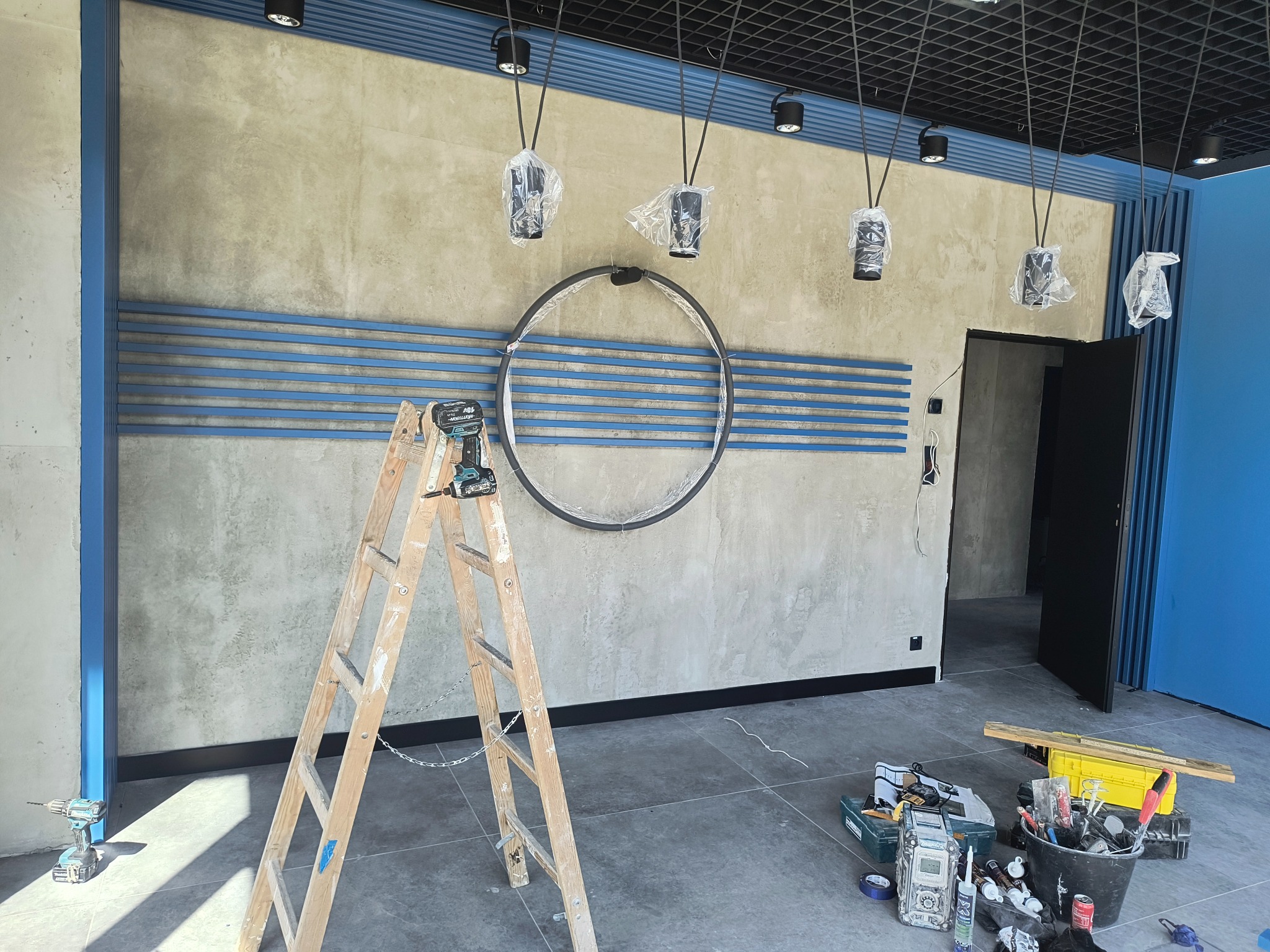 Ściana biura w trakcie wykańczania z dekoracyjnymi, poziomymi, niebieskimi listwami, okrągłą dekoracją, drabina z wiertarką i narzędzia na podłodze.