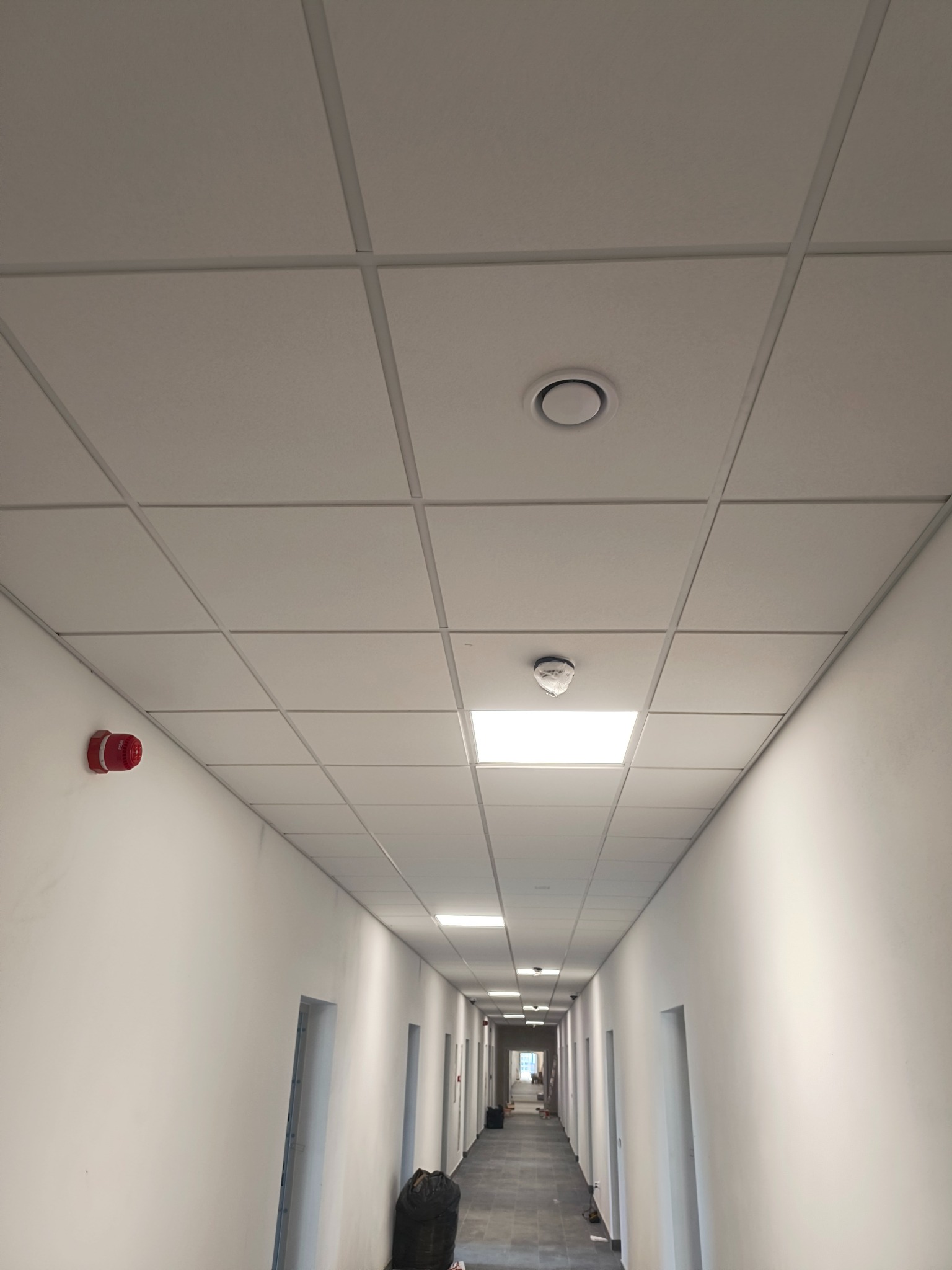Długi korytarz biurowy w trakcie wykańczania, widoczne białe ściany, szara podłoga, sufit kasetonowy z oświetleniem i czujnikiem dymu, czerwony alarm pożarowy na ścianie.