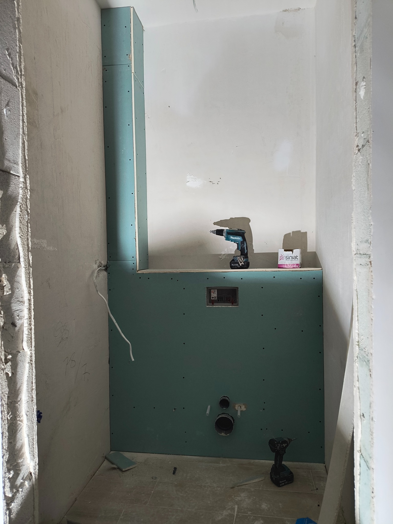 Wnętrze łazienki w trakcie zabudowy: zielone płyty na stelażu, widoczne otwory na instalacje, wkrętarka akumulatorowa Makita i opakowanie wkrętów na półce, surowe ściany w tle.