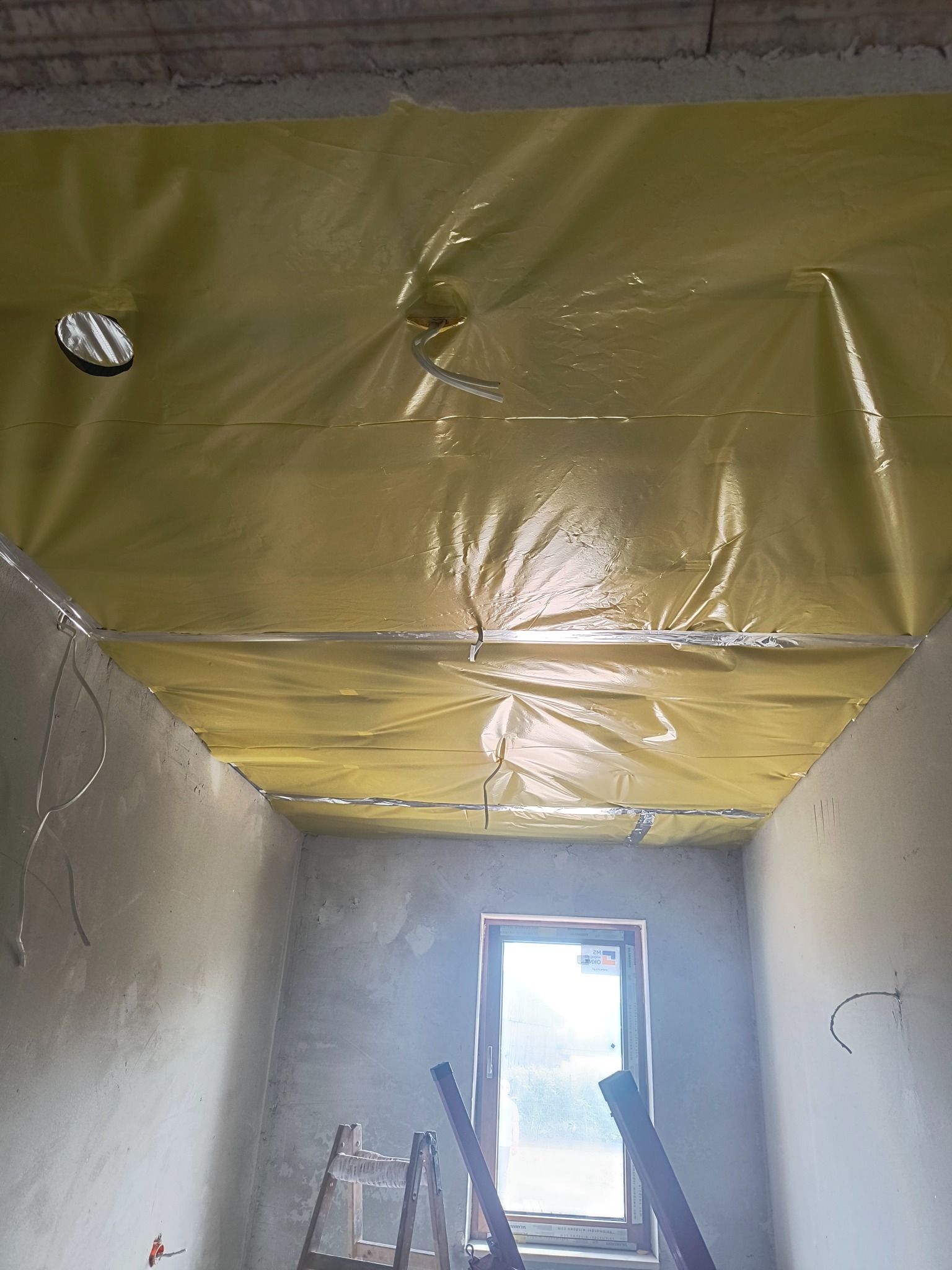 Pokój w trakcie prac wykończeniowych: surowy tynk na ścianach, zabezpieczony folią sufit z wystającymi przewodami, drabina oparta o ścianę pod oknem.