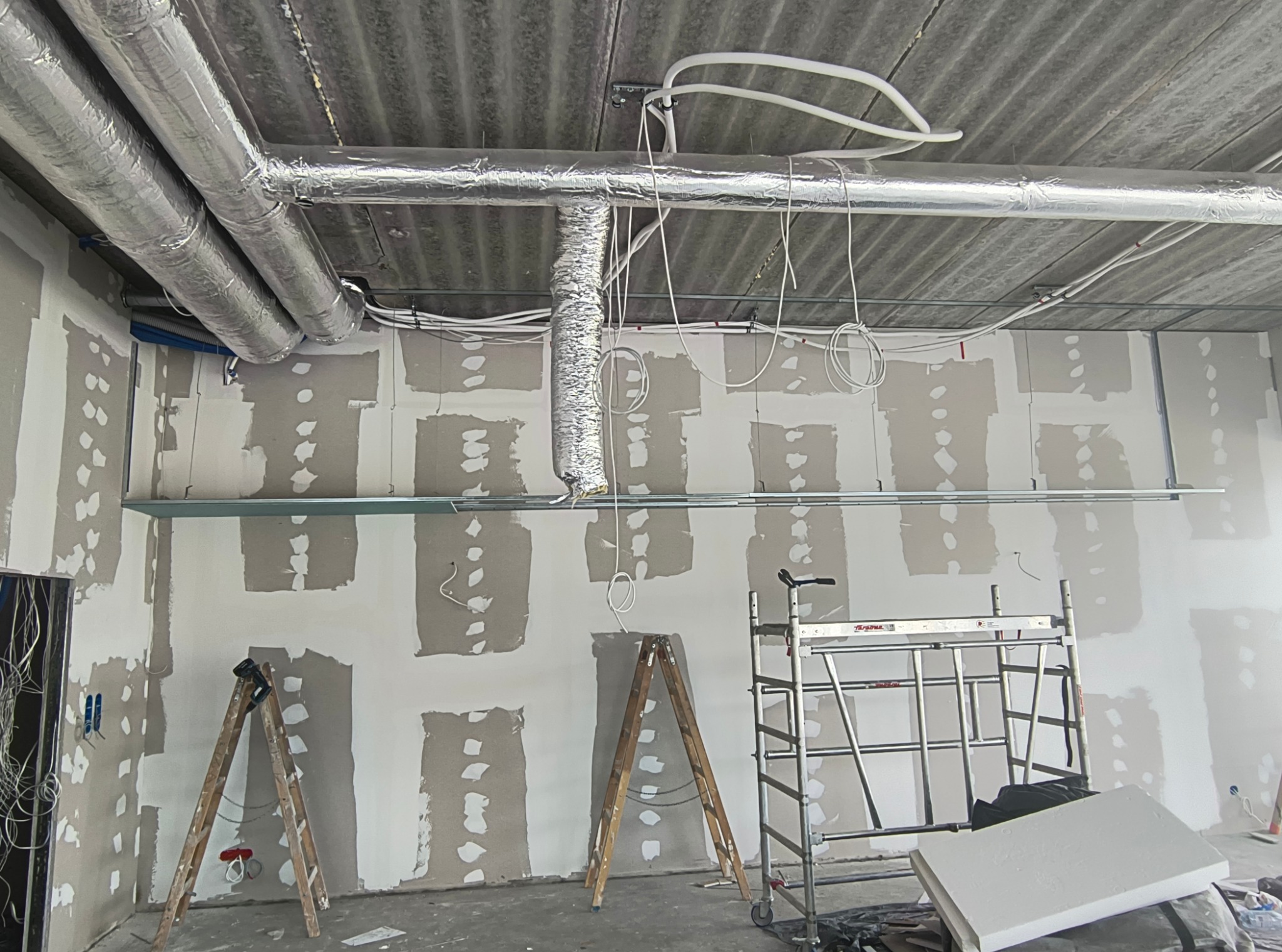 Wnętrze w trakcie prac wykończeniowych: ściany z płyt karton-gips, częściowo zaszpachlowane, widoczne rury wentylacyjne pod sufitem, drabiny i rusztowanie.