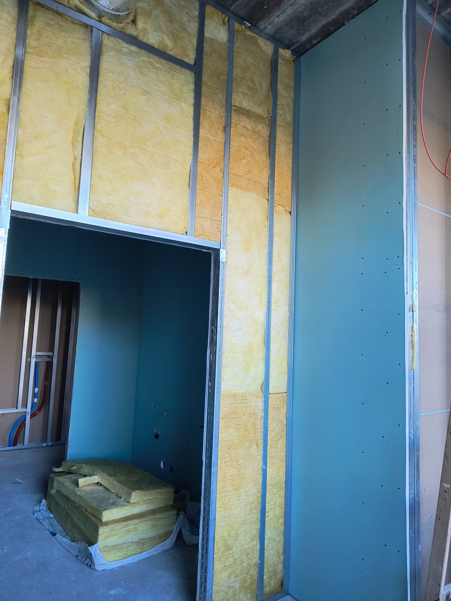 Wnętrze w trakcie prac wykończeniowych: metalowy stelaż, wełna mineralna w ścianie, częściowo zamontowana płyta na ścianie, widoczne przewody instalacji.
