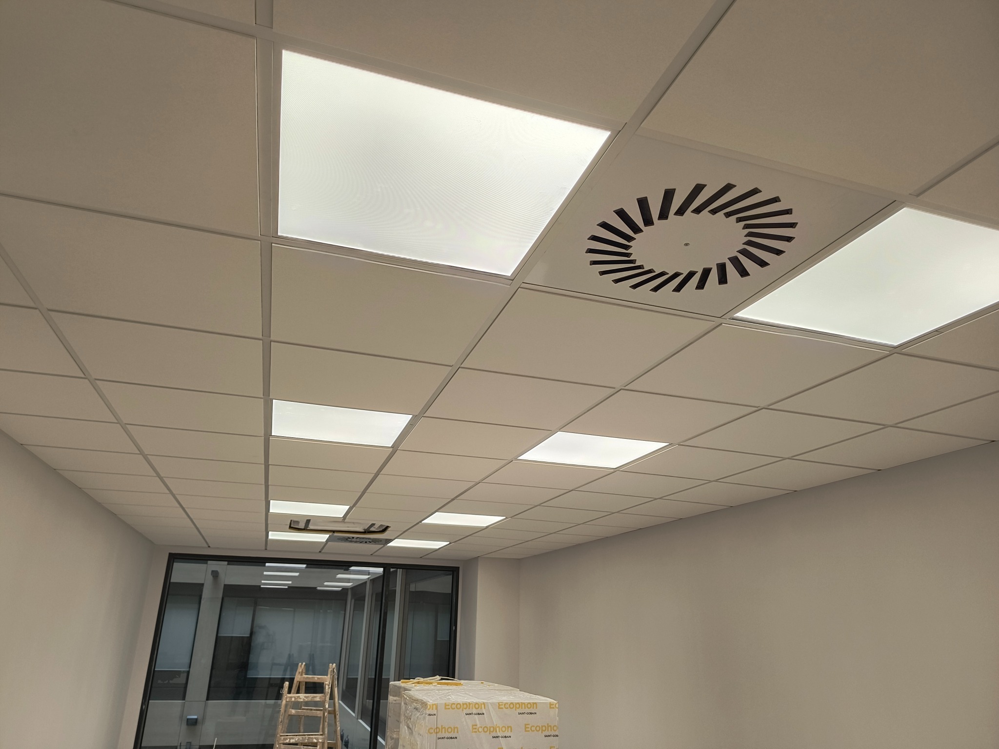 Kasetonowy sufit podwieszany z wbudowanym oświetleniem i okrągłym nawiewnikiem wentylacyjnym, widoczne materiały budowlane i drabina w tle.