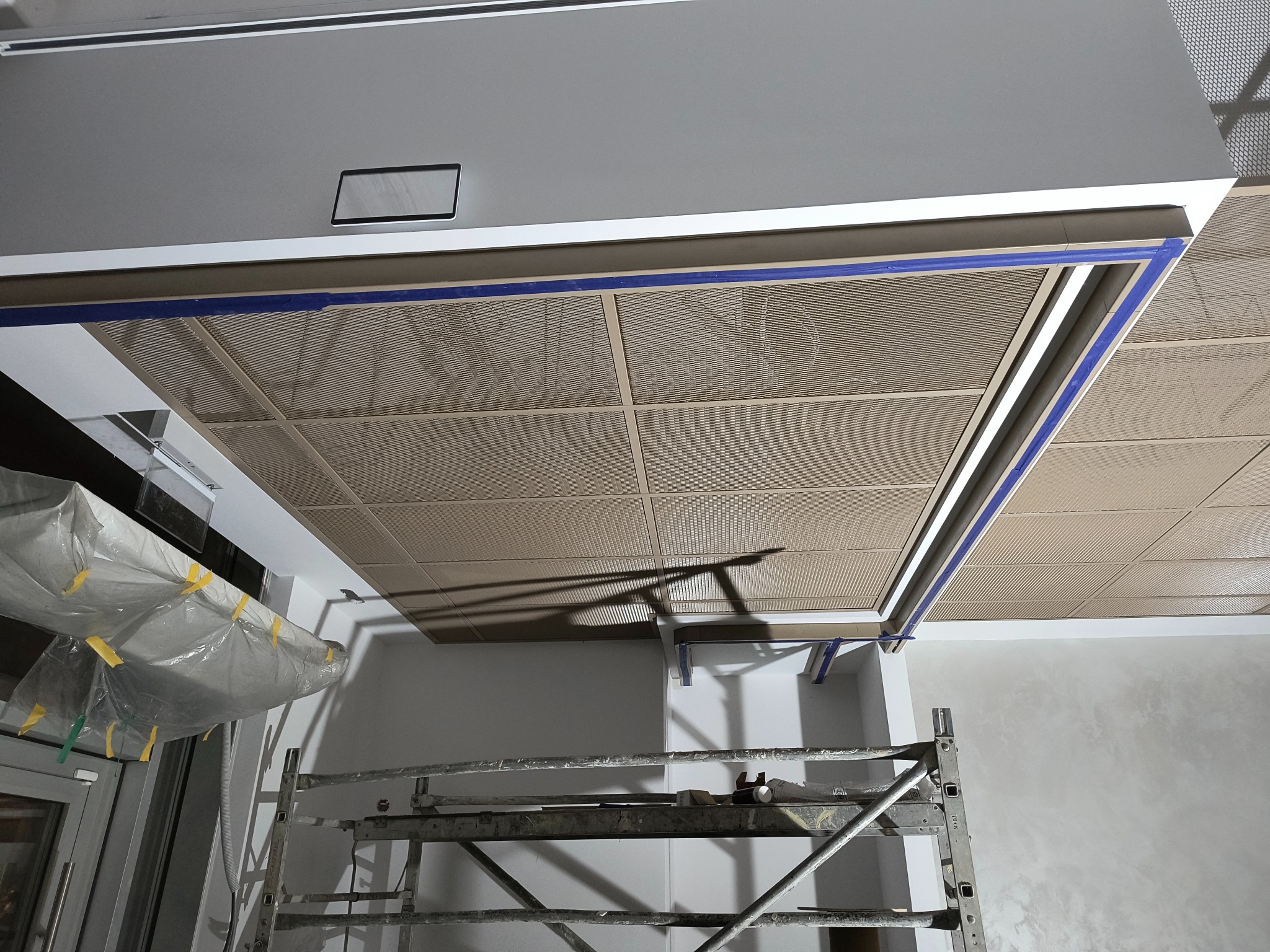 Montaż kasetonowego sufitu podwieszanego z widocznym rusztowaniem, zabezpieczoną folią okiennicą i niebieską taśmą malarską na obrzeżach sufitu.