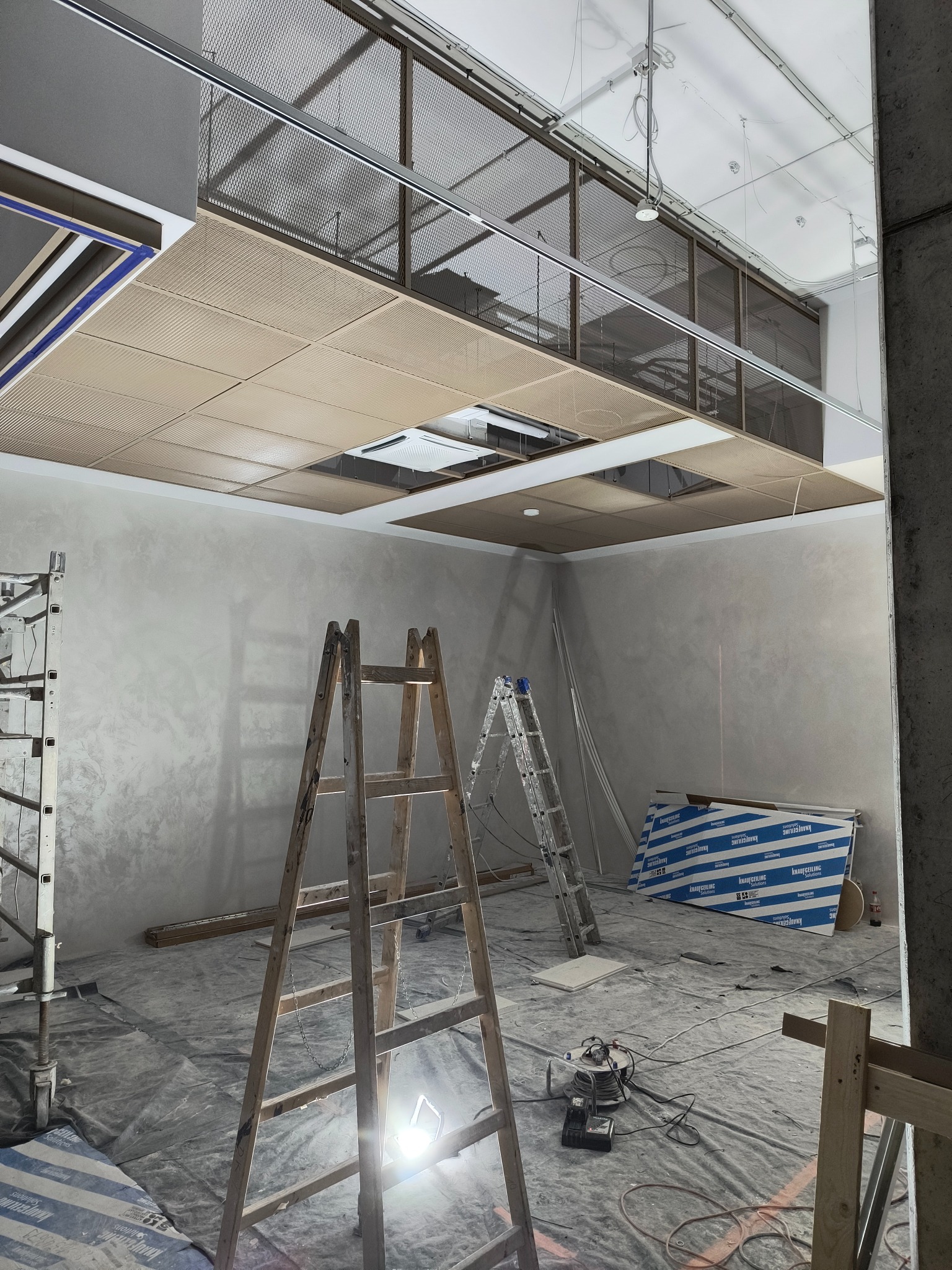 Montaż perforowanego sufitu podwieszanego w pomieszczeniu biurowym, widoczne drabiny, narzędzia i zabezpieczona folią podłoga.