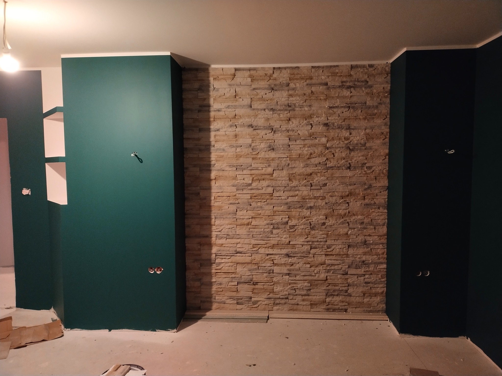 Ściana wykończona dekoracyjną okładziną kamienną, sąsiadująca z pomalowanymi na ciemnozielono ścianami w surowym stanie, widoczne gniazdka elektryczne i niedokończona podłoga.