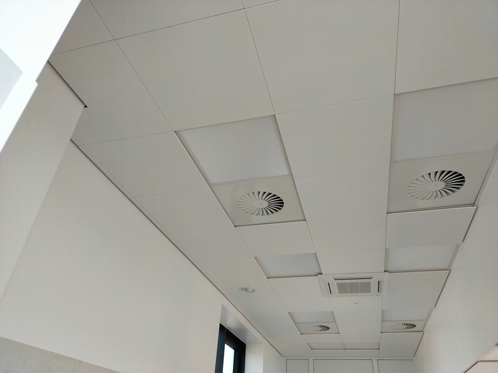 Kwadratowy sufit podwieszany z wbudowanymi panelami oświetleniowymi, kratkami wentylacyjnymi i klimatyzacją, widziany pod kątem.