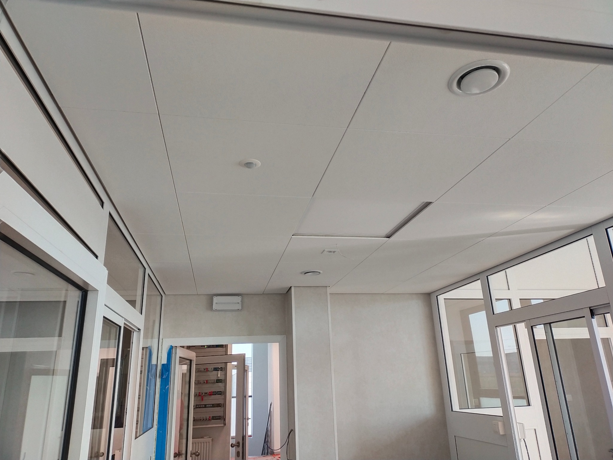 Biały sufit podwieszany z panelami rastrowymi, kratką wentylacyjną i oświetleniem w biurze w Kamionce, widoczne szklane drzwi i ściany.