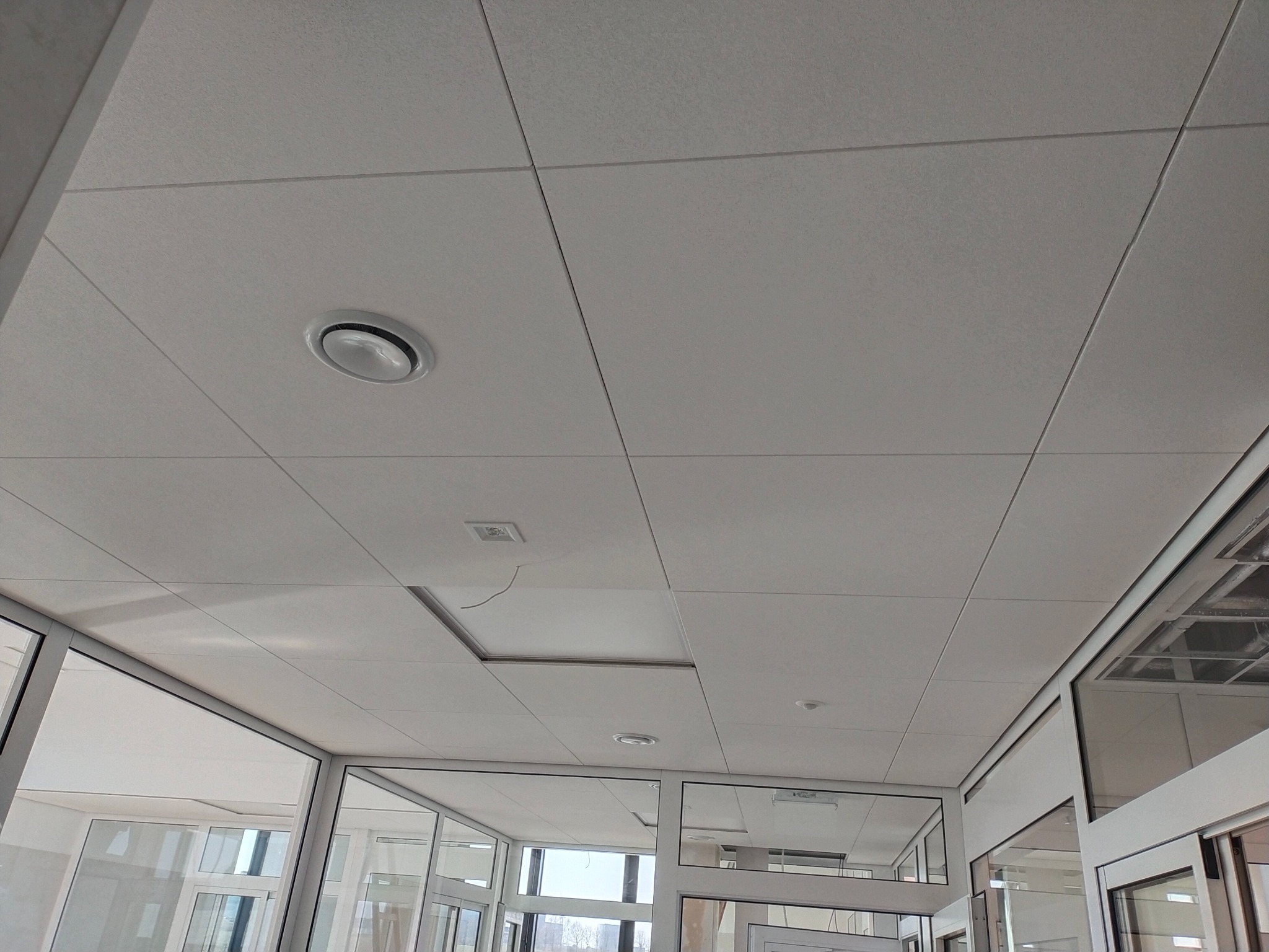 Biały, kasetonowy sufit podwieszany z wbudowanym oświetleniem i widocznym panelem rewizyjnym, otoczony szklanymi ścianami działowymi.
