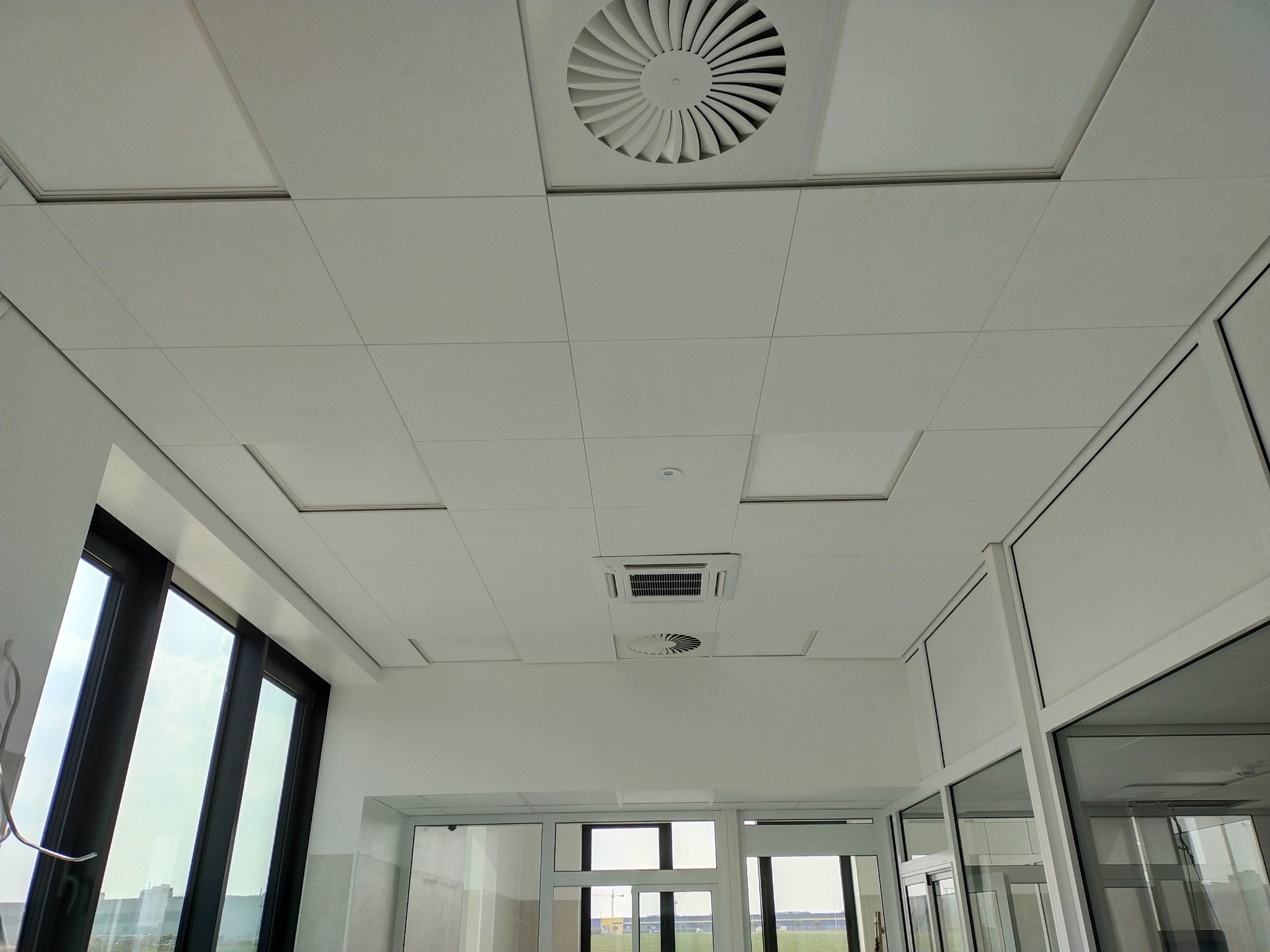 Biały sufit podwieszany z widocznymi panelami, kratkami wentylacyjnymi i oknami z widokiem na zewnątrz.
