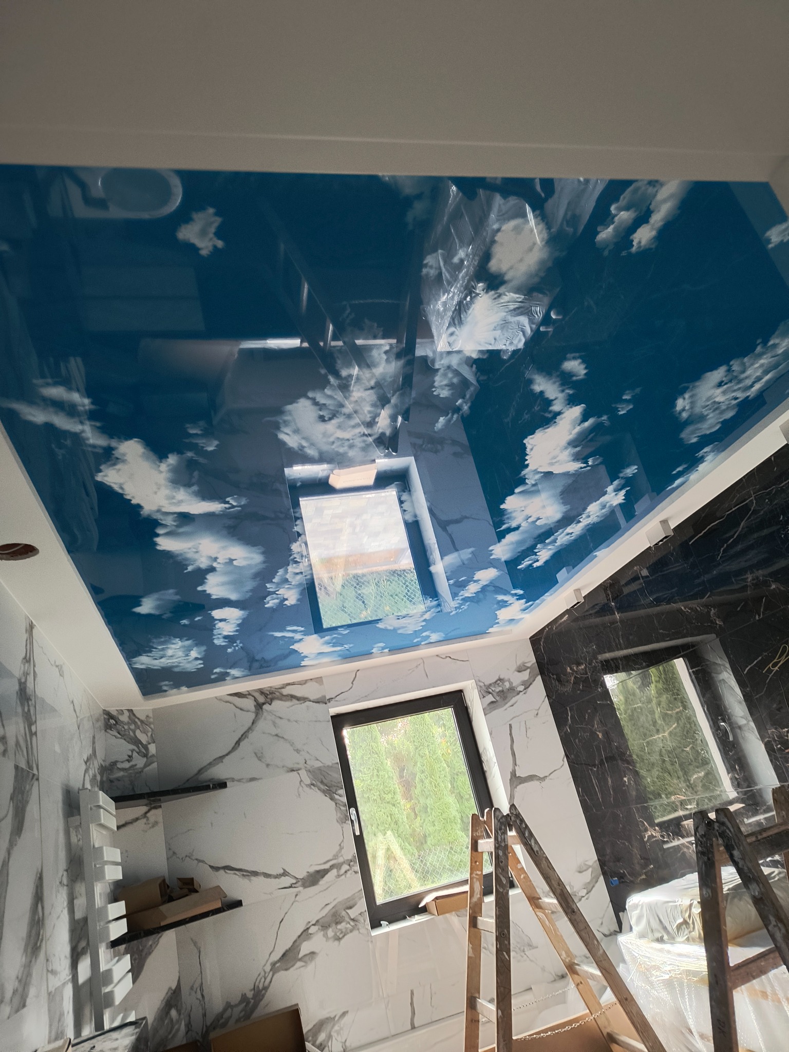 Błękitne, lustrzane wykończenie sufitu z motywem chmur w łazience z marmurowymi ścianami, widoczne okno i drabina malarska.