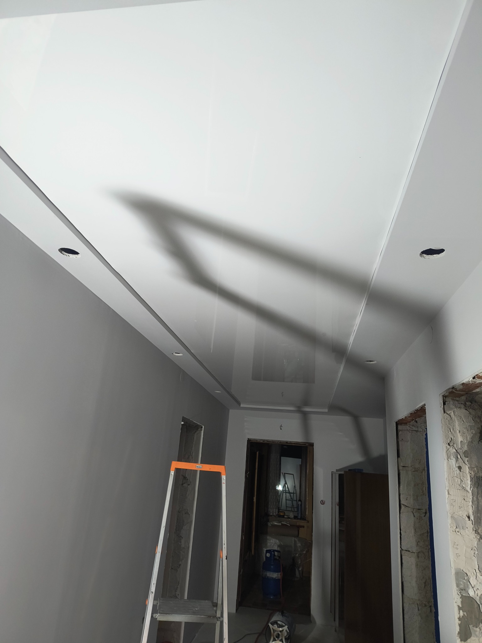 Biały, błyszczący sufit z wbudowanymi punktami oświetleniowymi w trakcie montażu, widoczna drabina i przygotowane otwory w ścianach.