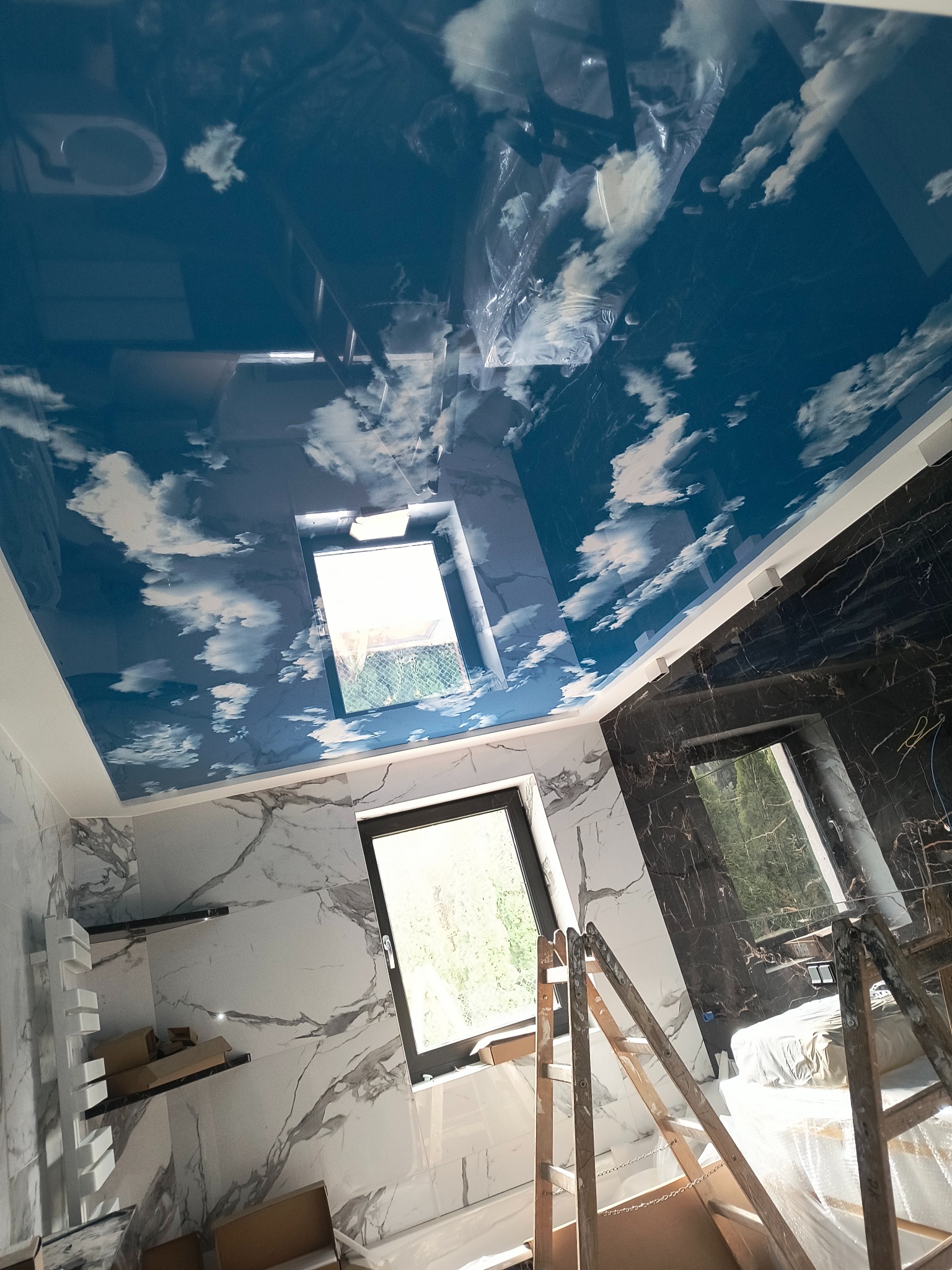 Błękitny, błyszczący sufit z motywem chmur odbija się w marmurowych płytkach ścian łazienki z oknami, widoczna drabina malarska.
