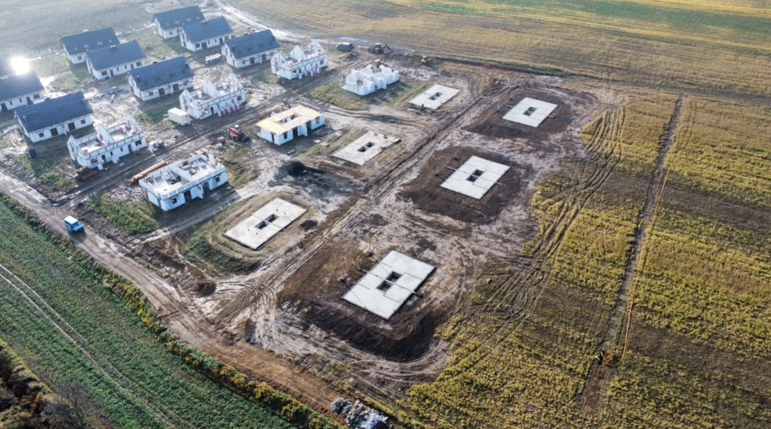 Widok z lotu ptaka na plac budowy osiedla domów jednorodzinnych w różnym stadium zaawansowania: od gotowych fundamentów z wykopami, przez domy w stanie surowym otwartym, po domy z dachem i oknami...