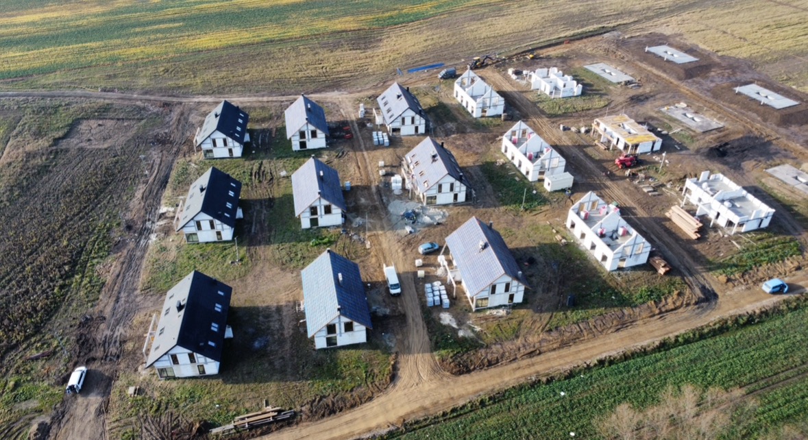 Widok z drona na osiedle w trakcie budowy: domy w różnym stadium zaawansowania, od fundamentów po konstrukcje z dachem, teren przygotowany pod kolejne budynki.