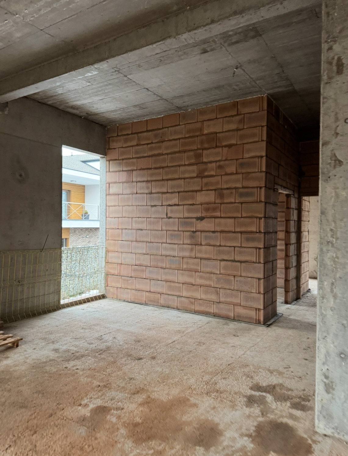 Wnętrze surowego budynku w trakcie budowy z widocznymi ścianami z pustaków ceramicznych, betonowym stropem i podłogą oraz oknem z widokiem na sąsiedni budynek.