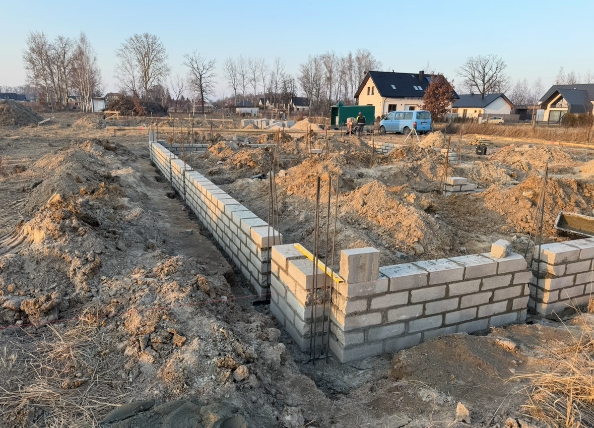 Wznoszenie muru fundamentowego z bloczków betonowych na placu budowy, widoczne stalowe pręty zbrojeniowe i poziomica. W tle zabudowa jednorodzinna.