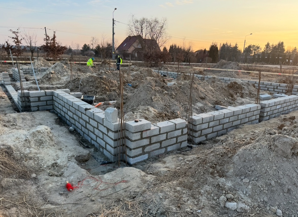 Wznoszenie ścian fundamentowych z bloczków betonowych na działce budowlanej, widoczni pracownicy w odblaskowych kamizelkach, w tle dom jednorodzinny i drzewa, poranek.