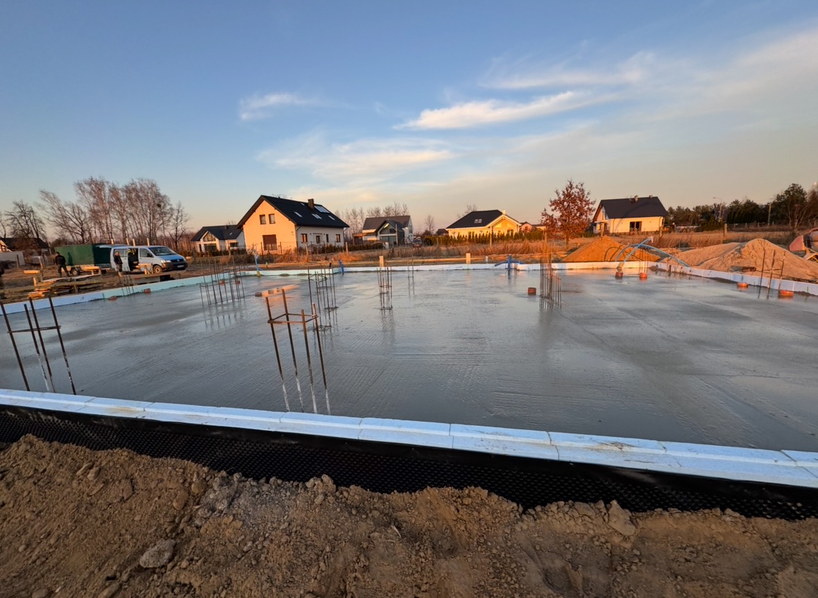 Wylana płyta fundamentowa z wystającymi prętami zbrojeniowymi, zabezpieczona izolacją pionową i poziomą, na tle zabudowy jednorodzinnej w promieniach zachodzącego słońca.