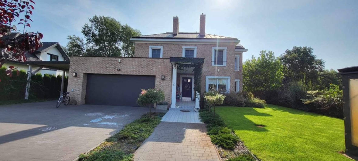 Dwukondygnacyjny dom z cegły klinkierowej, z garażem w bryle budynku, ciemną bramą garażową i wejściem z kolumnami oplecionymi zielenią, na zadbanym trawniku.