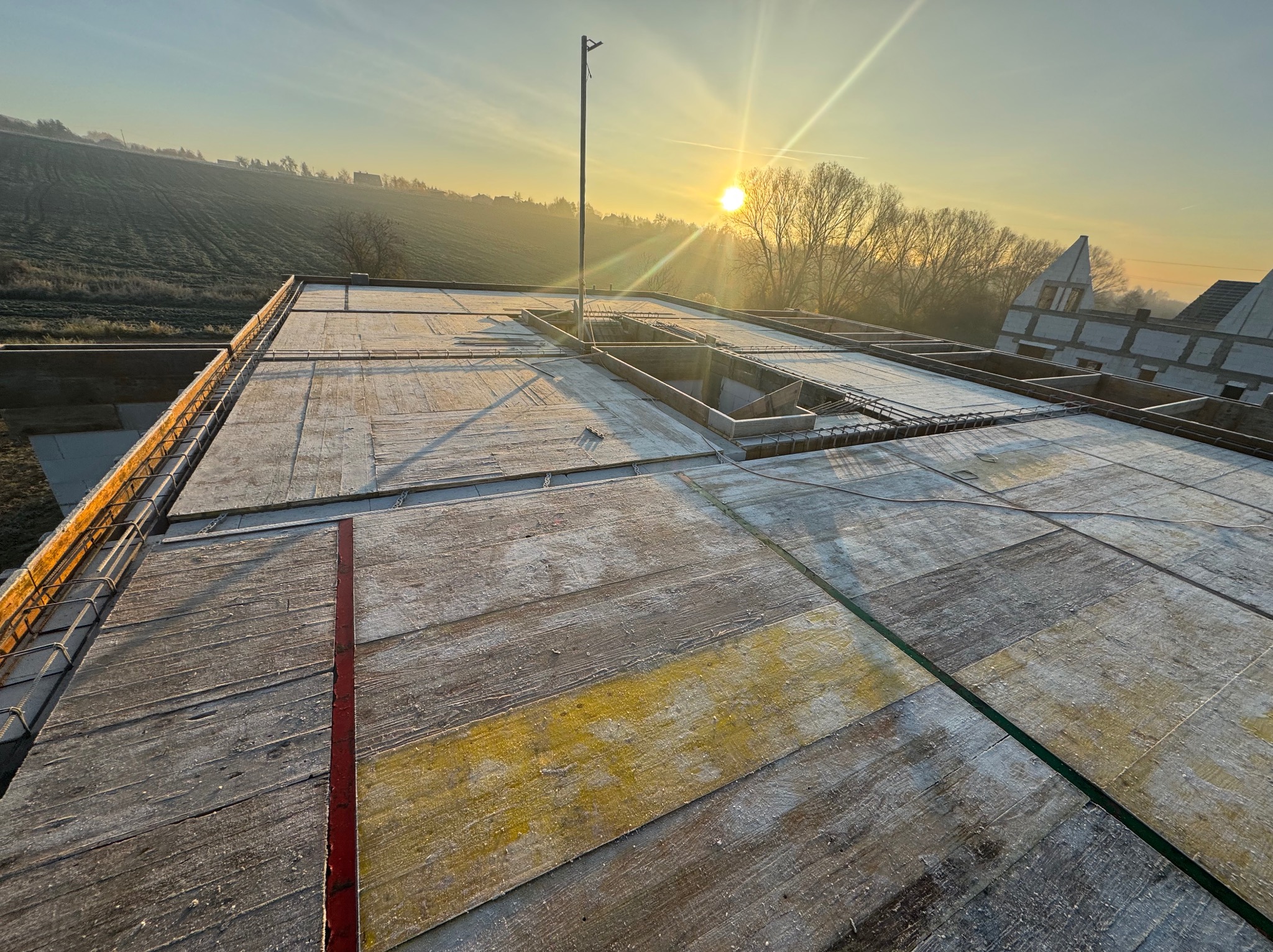 Zbrojenie stropu budynku w trakcie budowy, widok z góry na drewniane szalunki i stalowe pręty, w tle pagórkowaty krajobraz oświetlony wschodzącym słońcem.