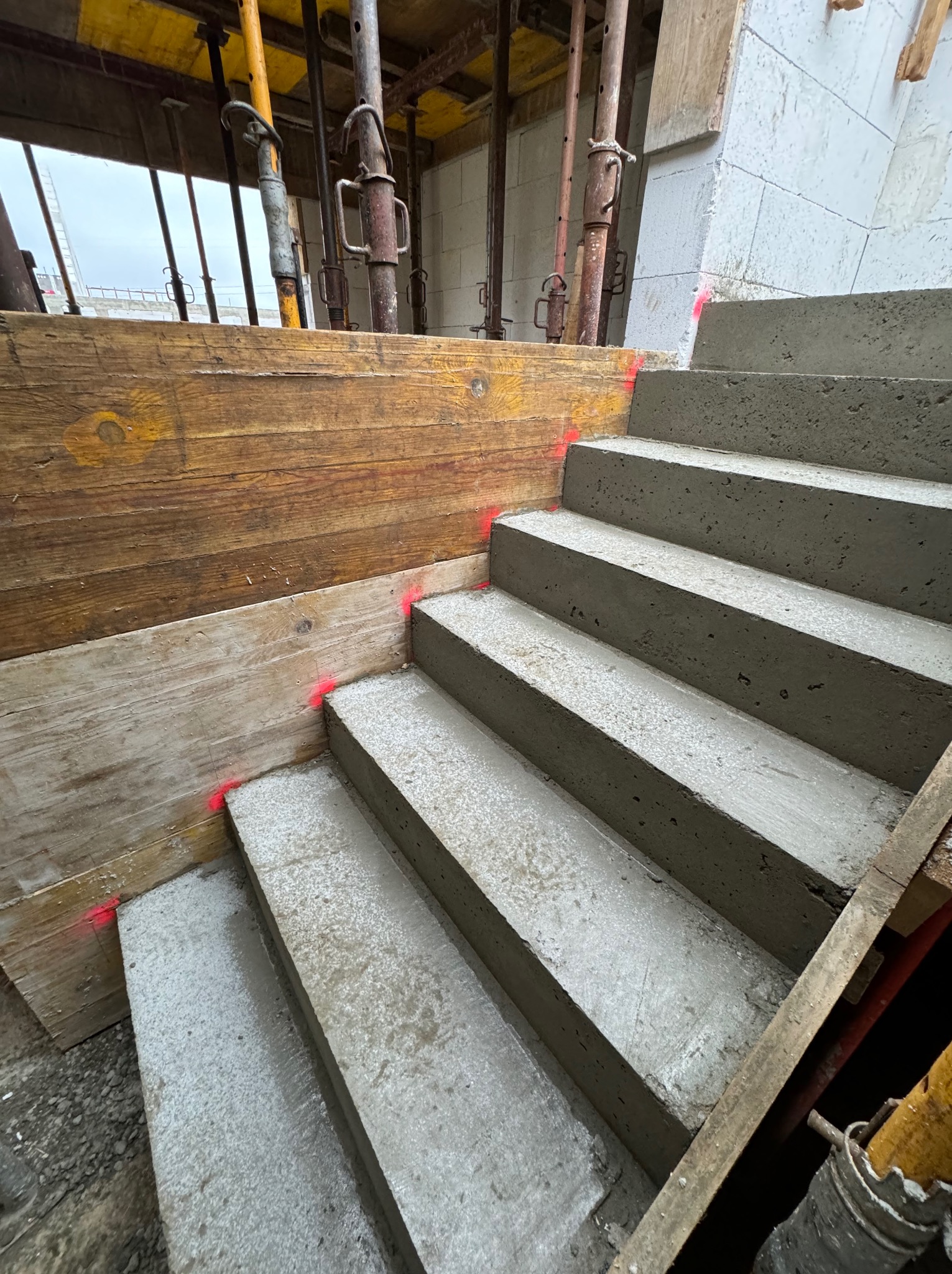 Betonowe schody w trakcie budowy, widoczne drewniane szalunki z czerwonymi oznaczeniami, podpory budowlane i fragment ściany z bloczków.