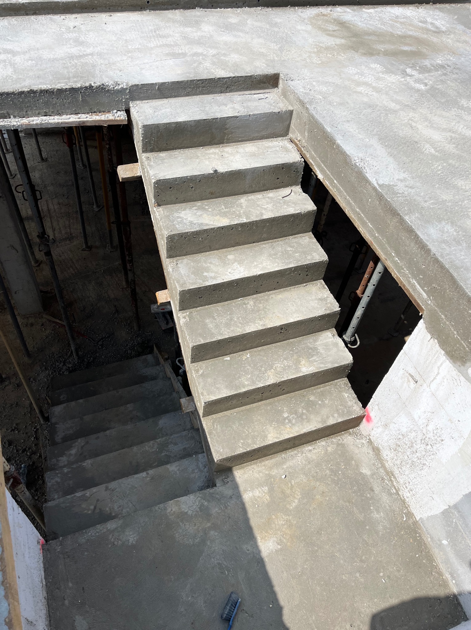 Surowe betonowe schody prowadzące w dół, widoczne stemple budowlane i drewniane szalunki, świeży beton.